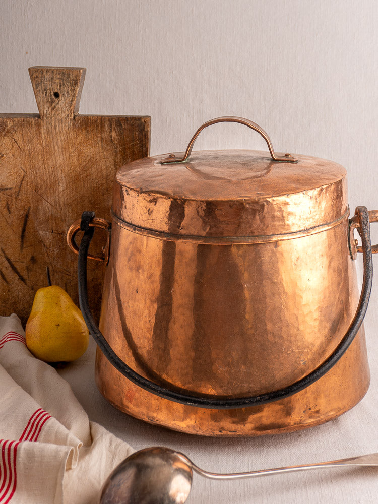 Antique French copper cauldron, hand-hammered with lid and black handle, styled on table with cutting board and pear.