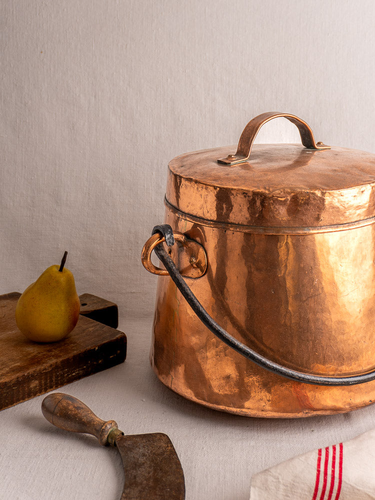 Antique French copper cauldron with lid next to cutting board, pear, metal knife, and striped white cloth on table.