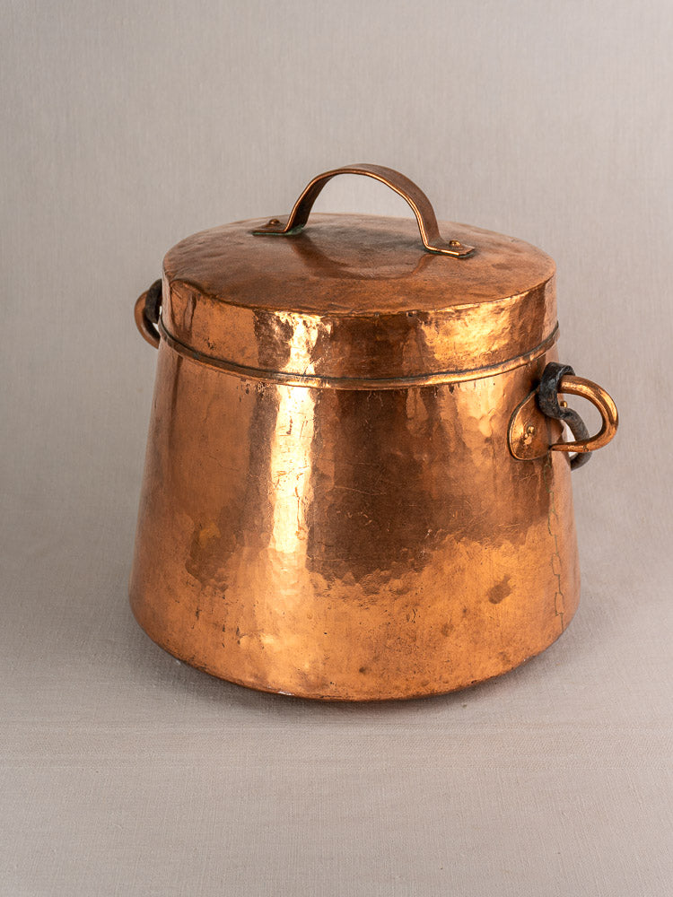 Hand-hammered antique French copper cauldron with lid and side handles on a light gray surface.