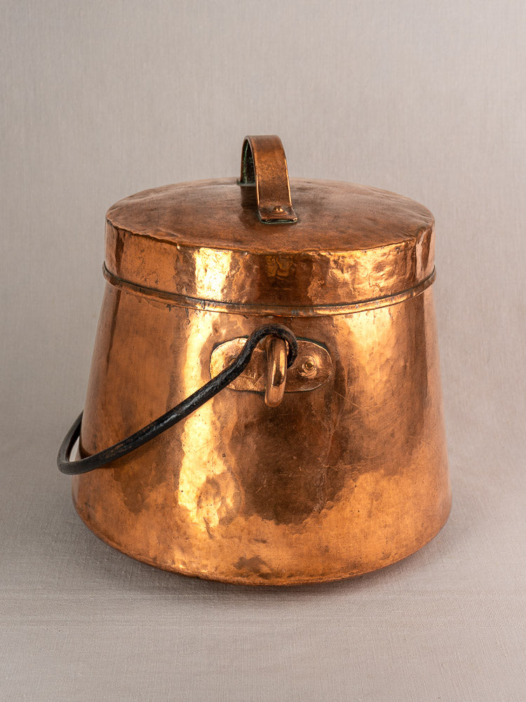 19th-century French hand-hammered copper lidded cauldron with curved side handle and warm patina, on a neutral gray background.
