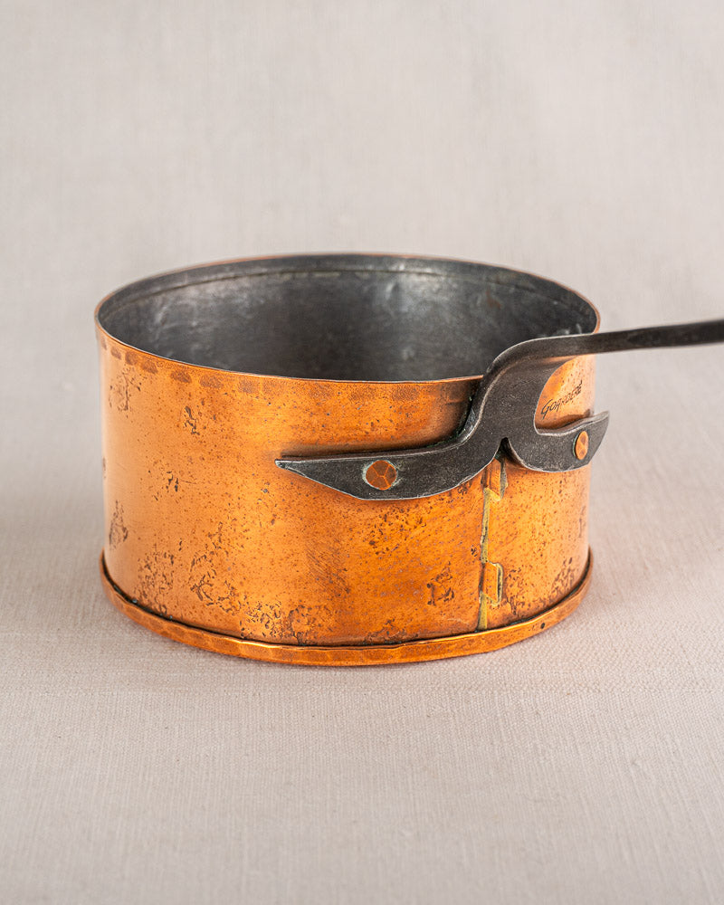 Round rustic copper pot with black interior and handle, set on a light beige surface.