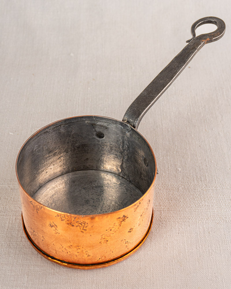 Small round copper French saucepan with long black handle, shown on a light textured surface.