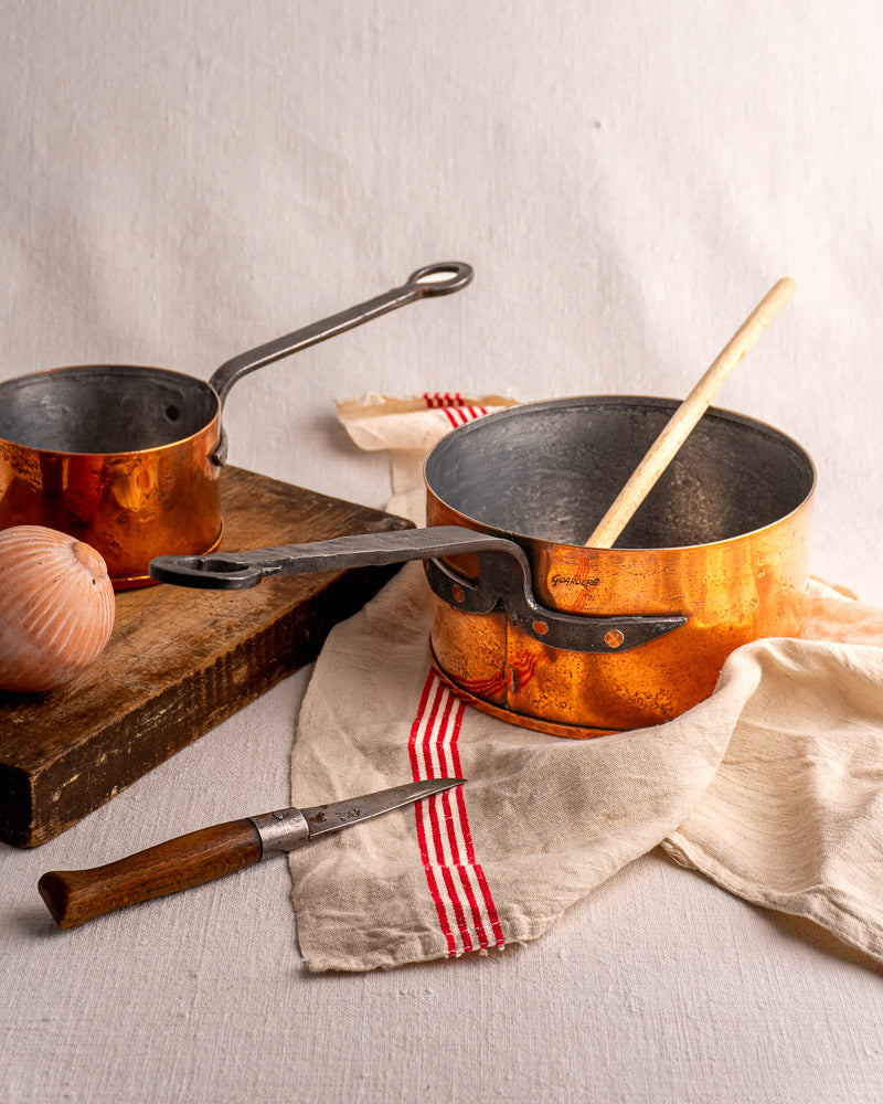 Jean Goardère vintage French copper saucepans with wooden spoon on striped cloth, onion and knife nearby.