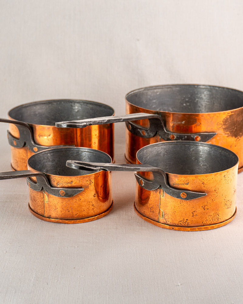 Set of four vintage Jean Goardère French copper saucepans with metal handles on light surface.