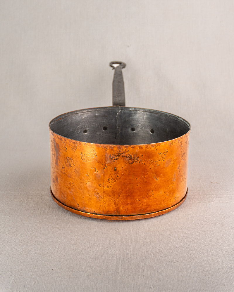 Tall, round copper saucepan with metal handle and holes near rim, on neutral surface.