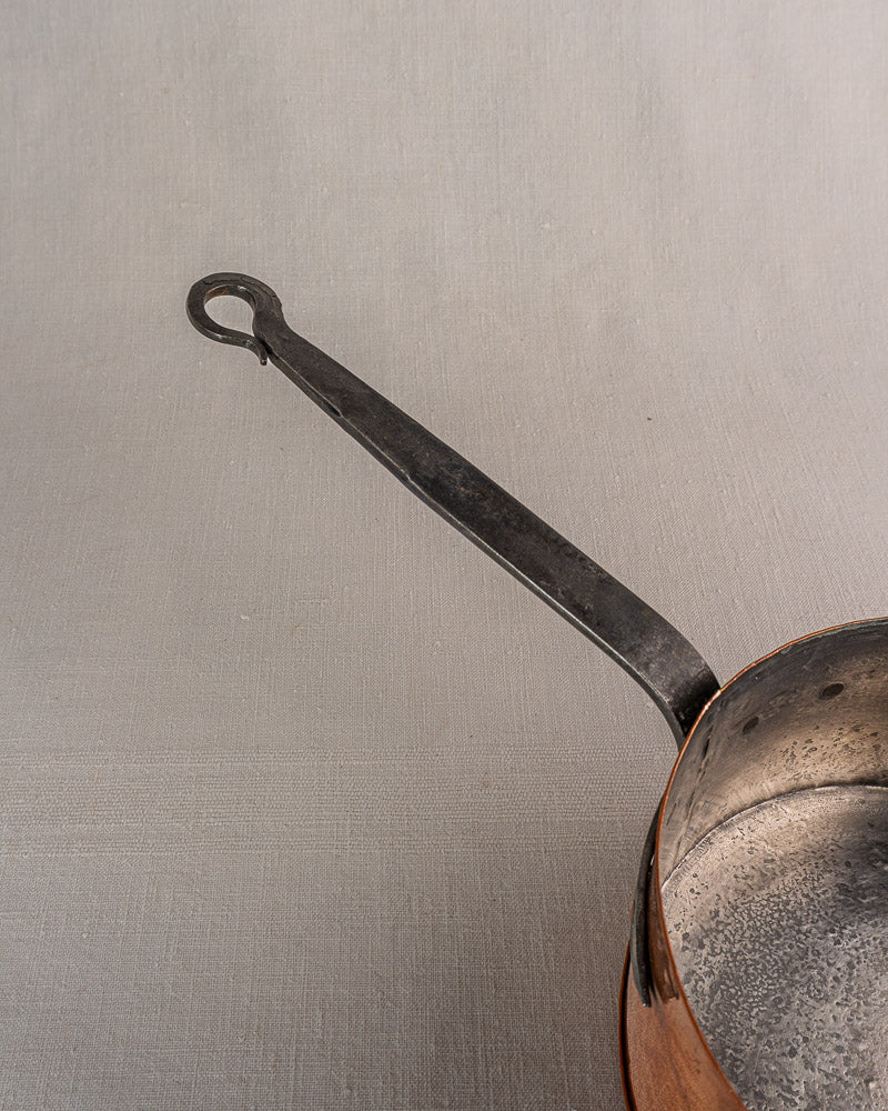 Vintage French copper saucepan with long black handle, worn interior, on light gray surface.