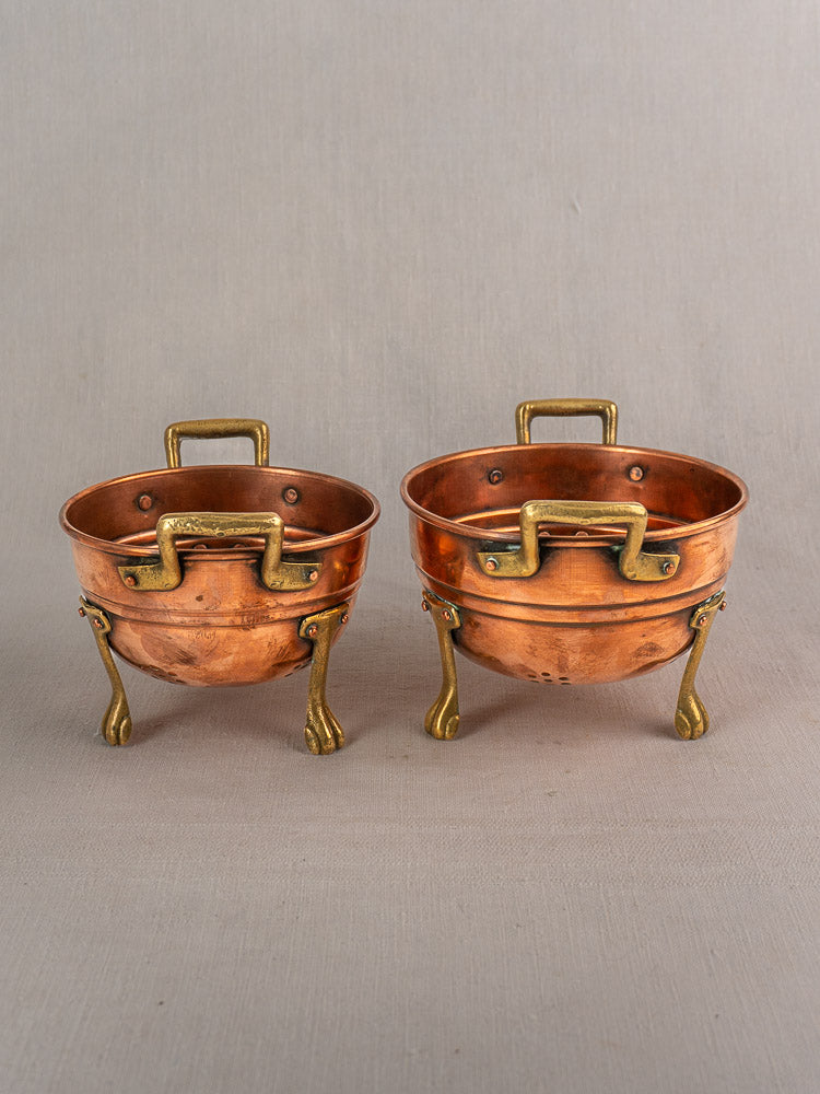 Three antique French copper colanders with brass paw feet, early 20th century, on neutral fabric.