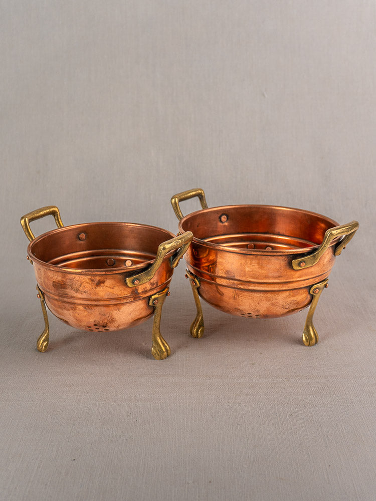 Set of three early 20th-century French antique copper colanders with brass paw feet on neutral fabric surface.