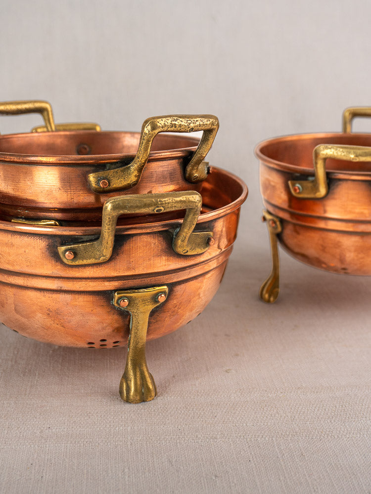 Antique French copper colander set of three with brass paw feet, stacked on a light beige surface.