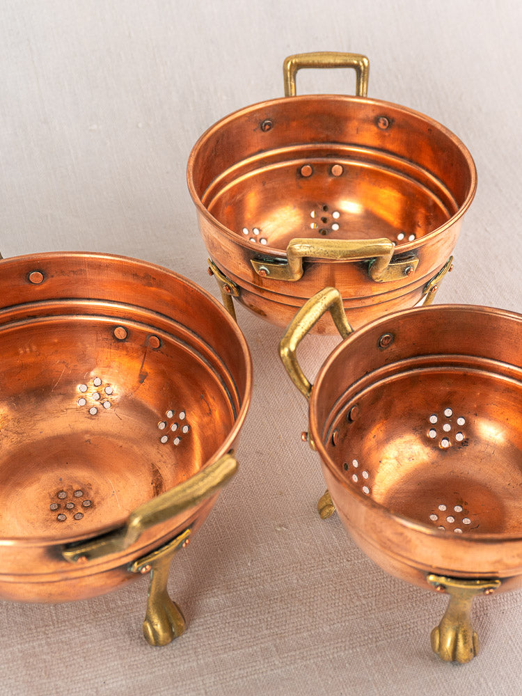 Antique French copper colander set of three with brass paw feet and decorative drainage holes, early 20th century.