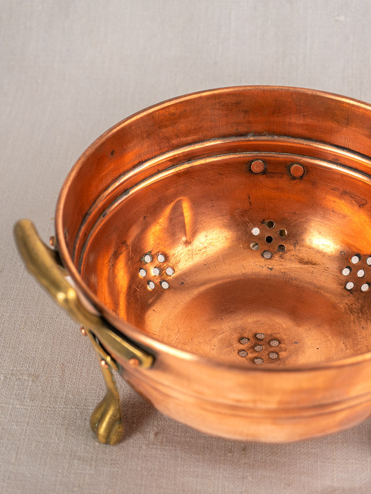 Antique French copper colander set of three with brass paw feet and floral drainage holes on beige fabric.
