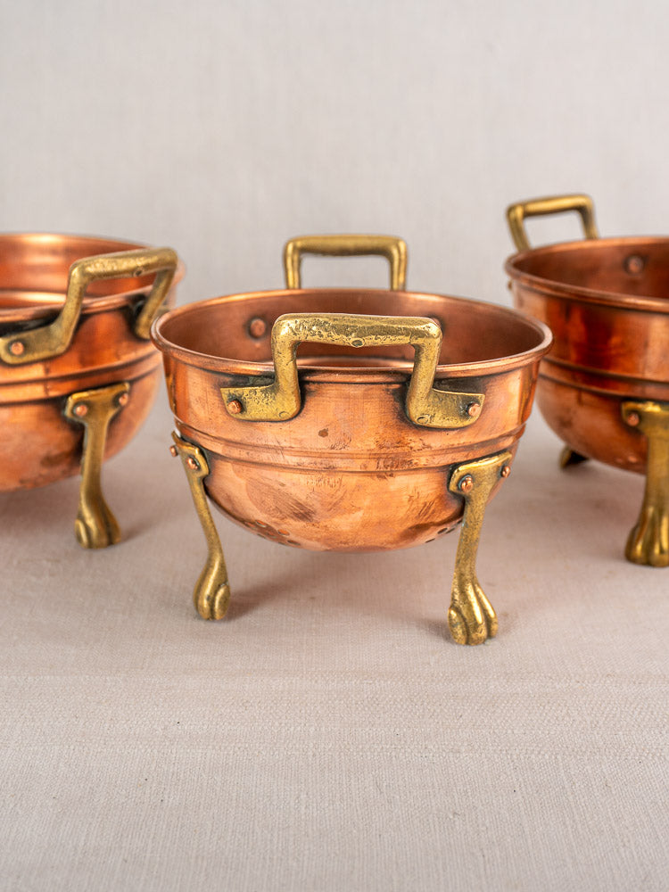 Set of three early 20th-century French copper colanders with brass paw feet and handles on light fabric.
