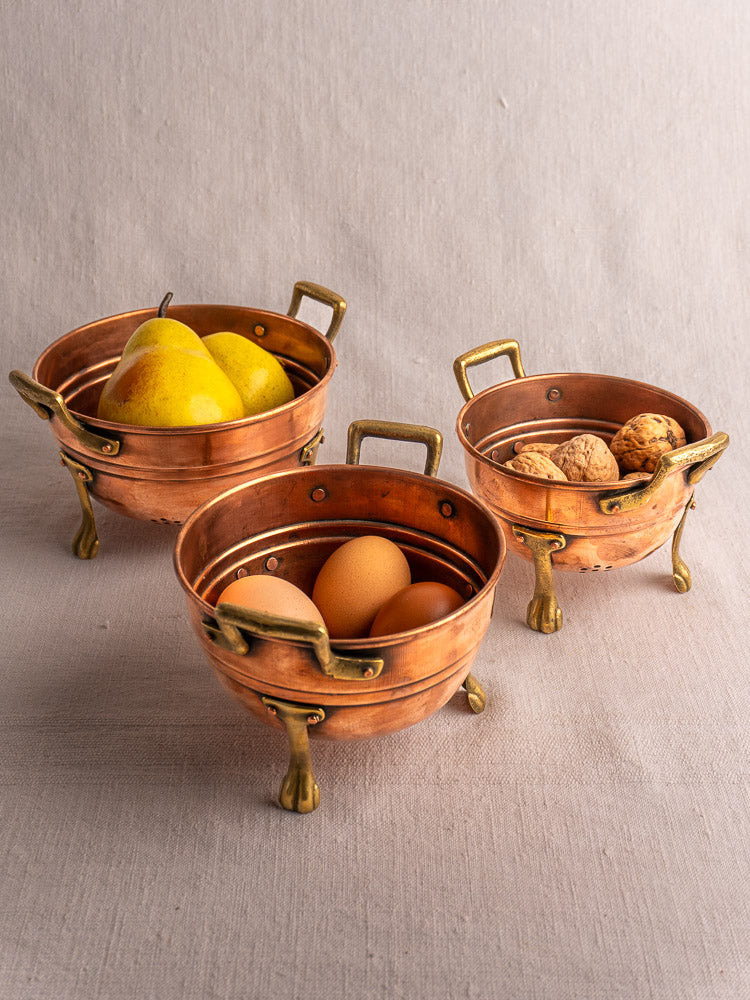 Set of three antique French copper colanders with brass paw feet, each filled with pears, eggs, or walnuts on beige fabric.