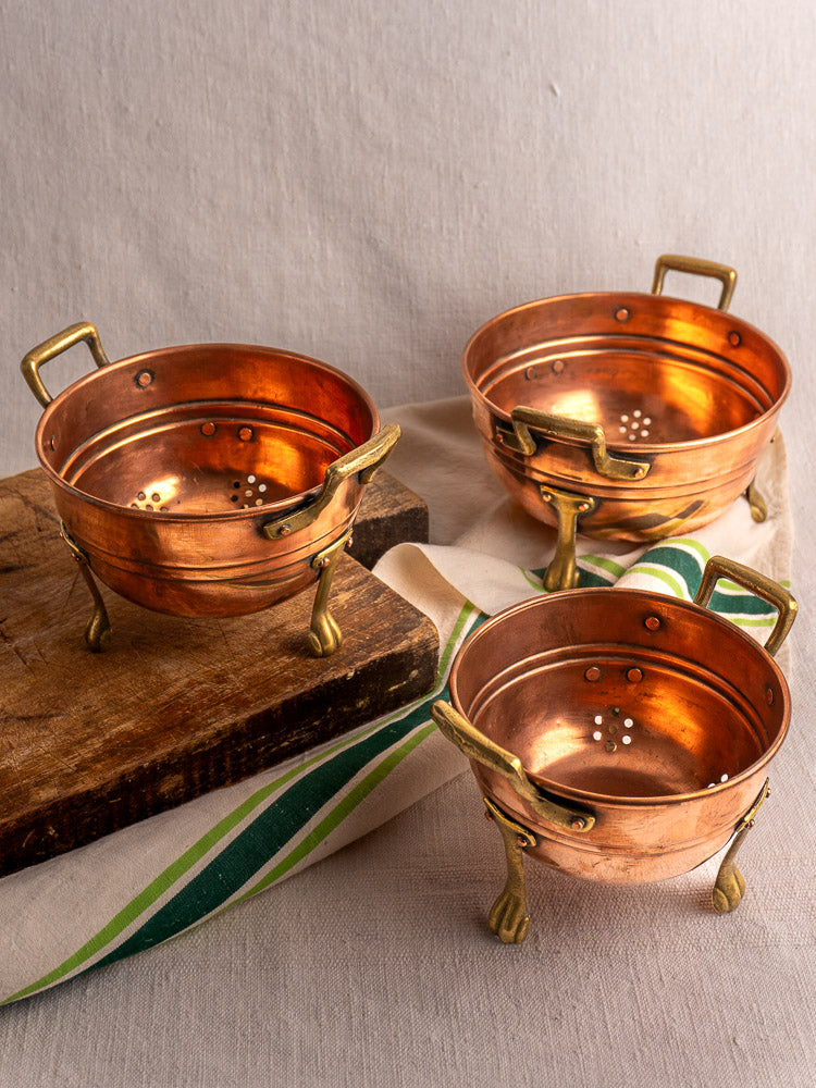 Set of three antique French copper colanders with brass paw feet on wood board and striped cloth.
