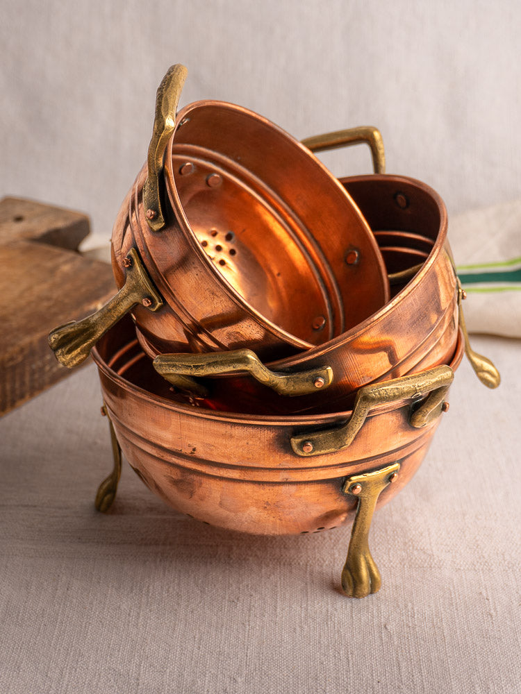 Antique French copper colander set of three with brass paw feet, stacked on light fabric.