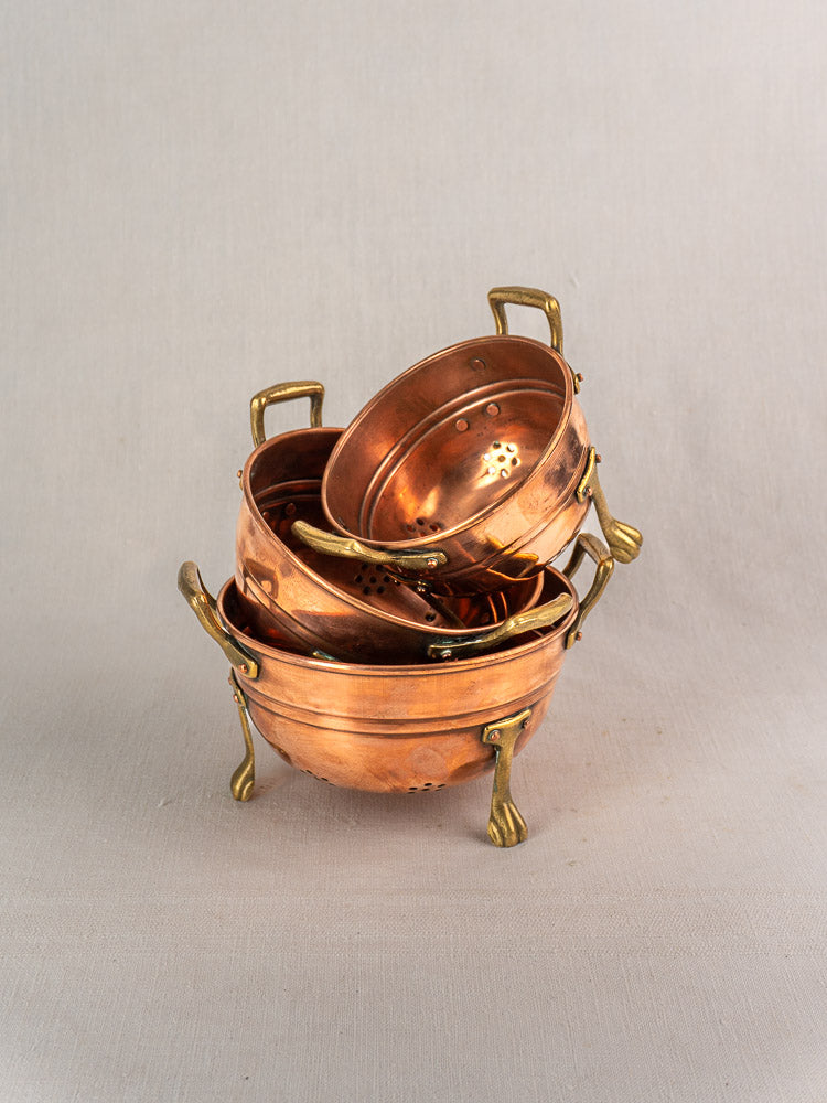 Set of three antique French copper colanders with brass paw feet, stacked on neutral fabric background.