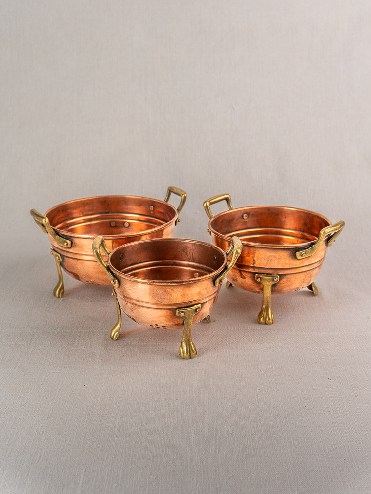 Set of three antique French copper colanders with brass paw feet, early 20th century, on neutral fabric.