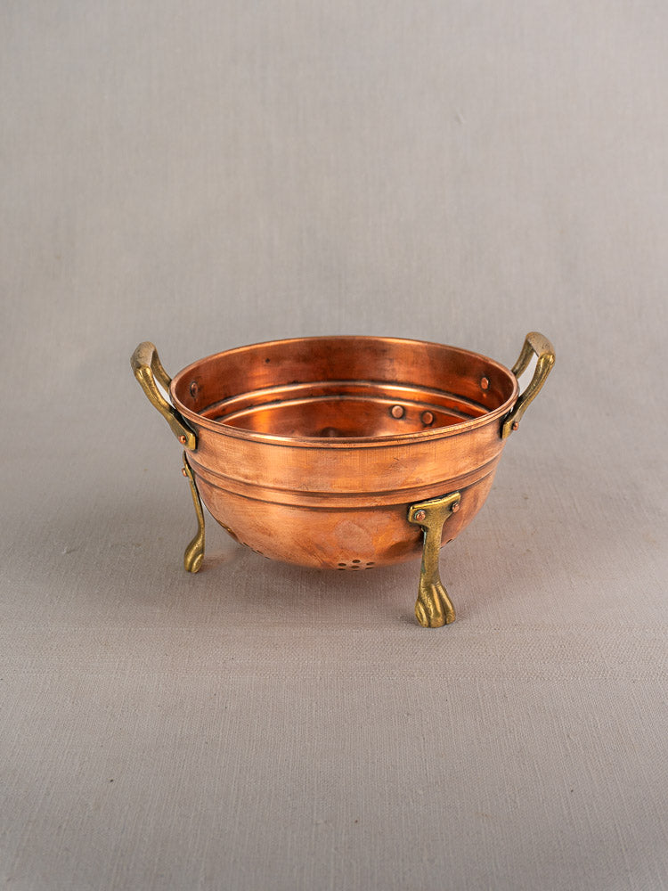 Set of three antique French copper colanders with brass paw feet and handles on a beige background.