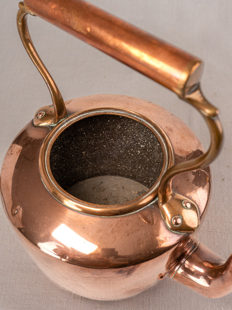 Antique French copper tea kettle with wood handle, open to show speckled interior, on neutral fabric.
