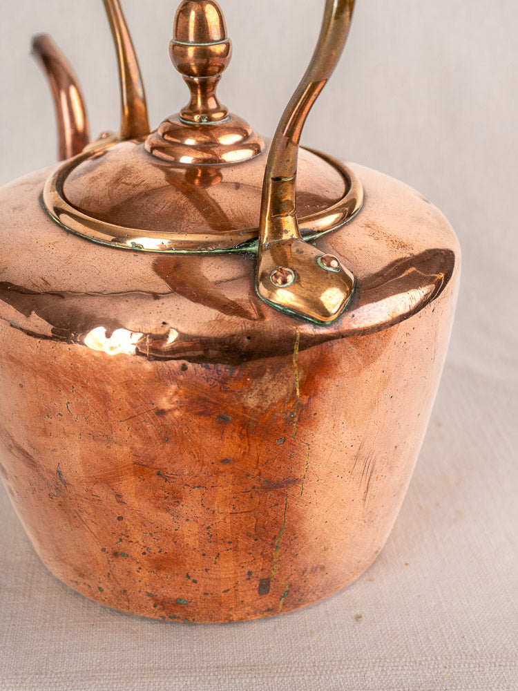Antique French copper tea kettle with cracked green-patinated body, gooseneck spout, and wood handle on fabric background.
