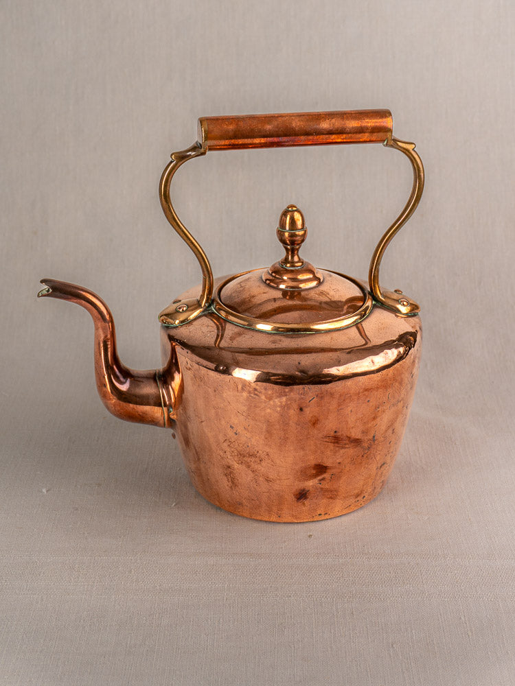 Antique French copper tea kettle with wood handle, curved spout, and brass acorn finial on neutral fabric.