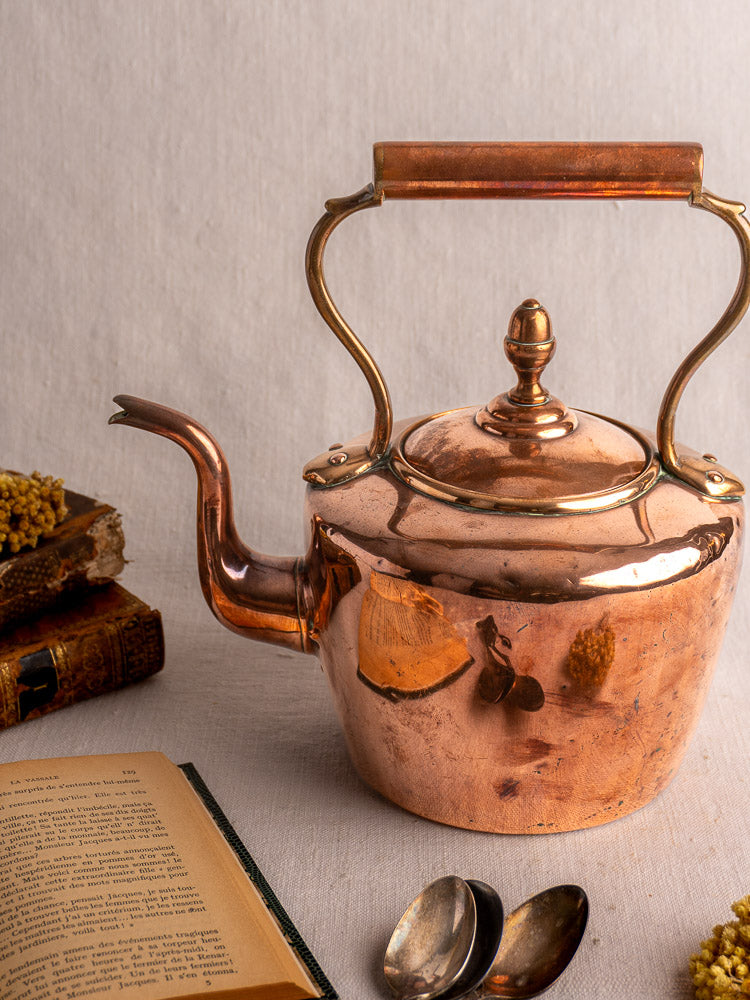 Antique French copper tea kettle with wood handle displayed beside vintage books, spoons, and yellow flowers on light fabric.