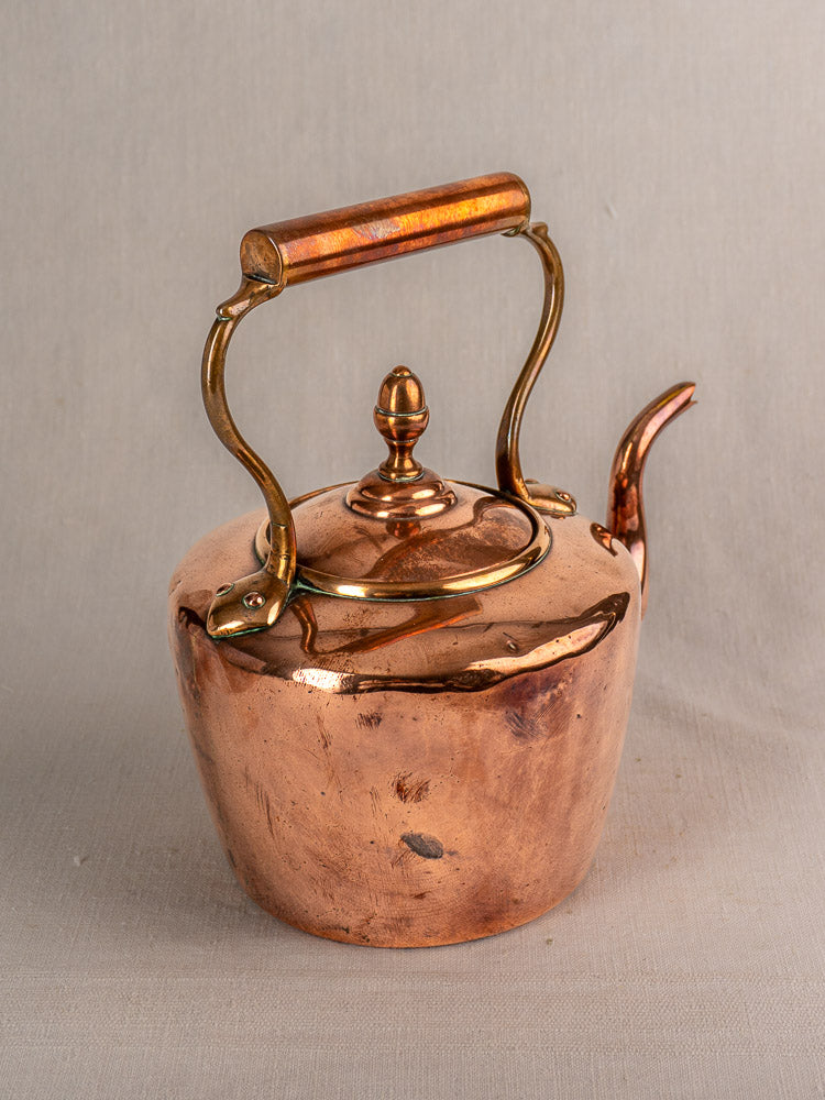 Antique French copper tea kettle with gooseneck spout, wood handle, and brass accents on neutral fabric background.