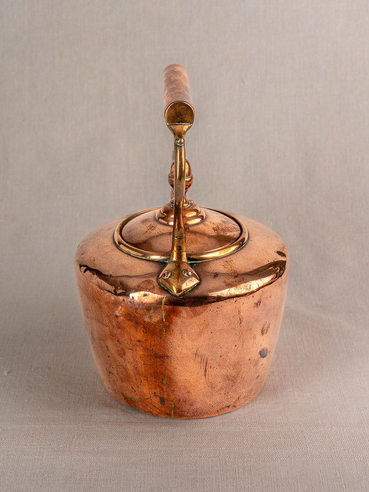 Antique French copper tea kettle with gooseneck spout, wood handle, acorn finial, on plain beige background.