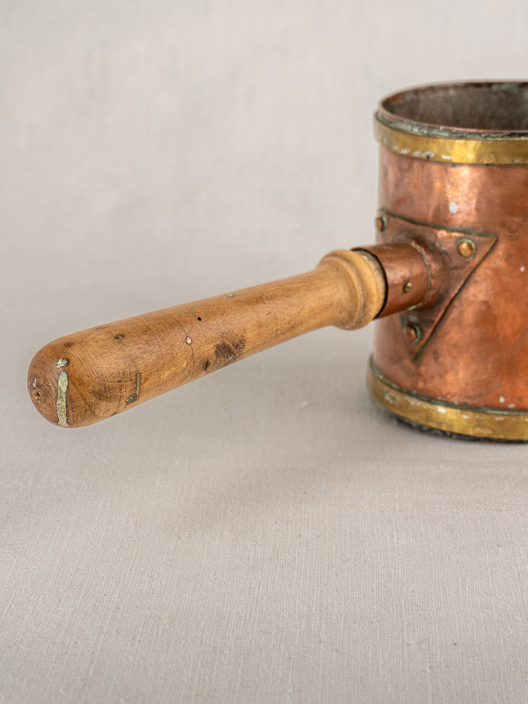 Antique French copper strainer pot with worn wooden handle and brass fittings on light beige fabric.