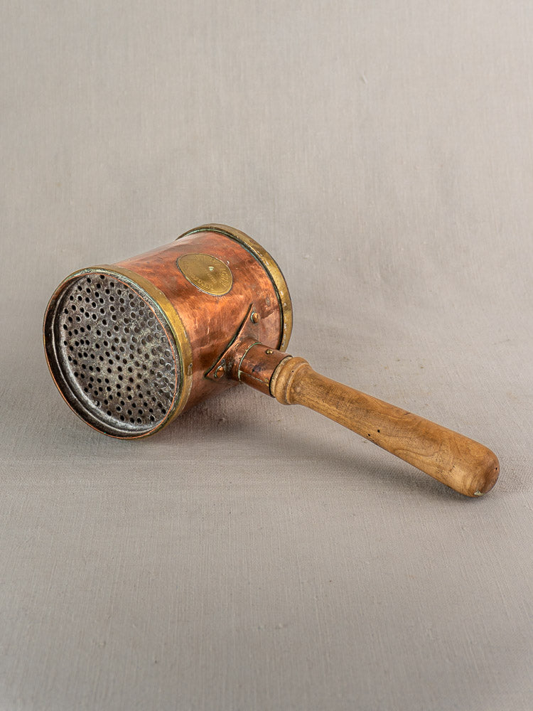 Antique French copper strainer pot with wooden handle, wall perforated, on neutral fabric background.