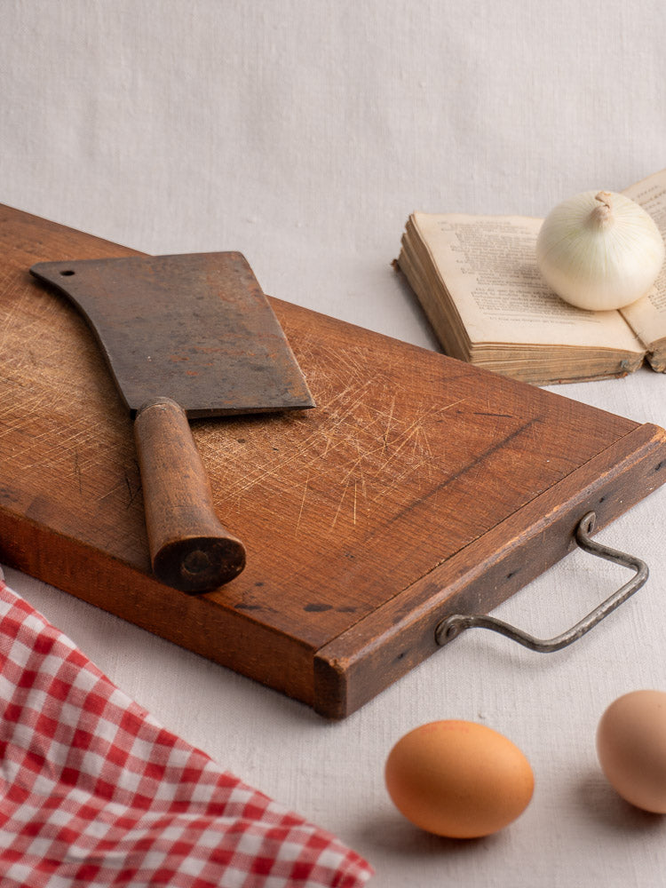 Antique French wood cutting board with iron handle, cleaver, cookbook, onion, eggs, and checkered cloth.