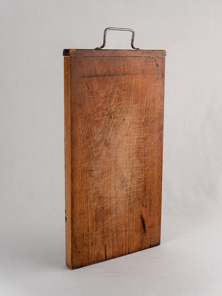Antique French wood cutting board with hand-forged iron handle, upright on a light surface against a plain background.