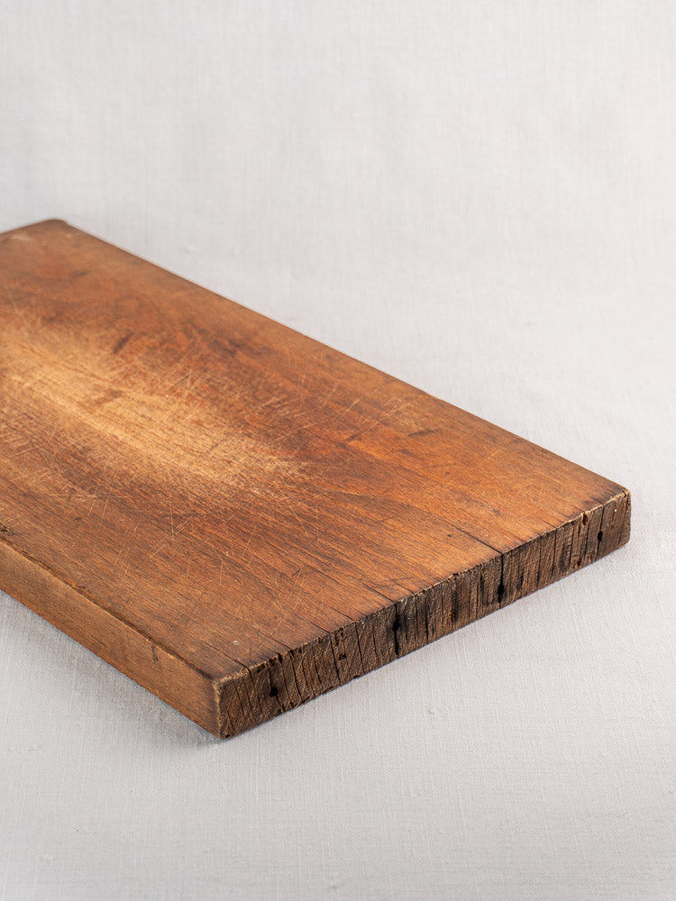 Antique French wood cutting board with iron handle, rough edges, and scratches on light fabric background.
