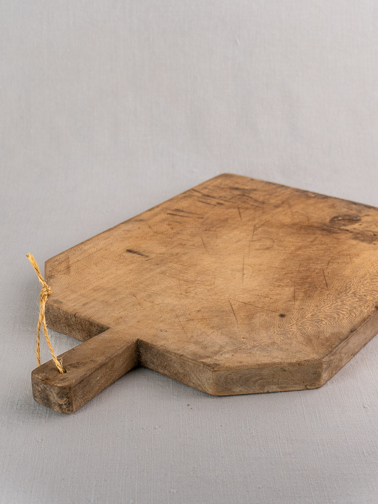 Antique French bread board in hand-carved beech with short handle and twine loop on light gray surface.
