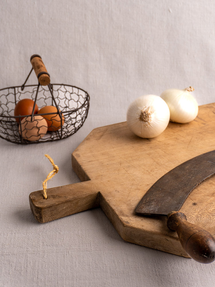 Antique French beech bread board with curved knife and onions, eggs in wire basket on gray fabric in background.