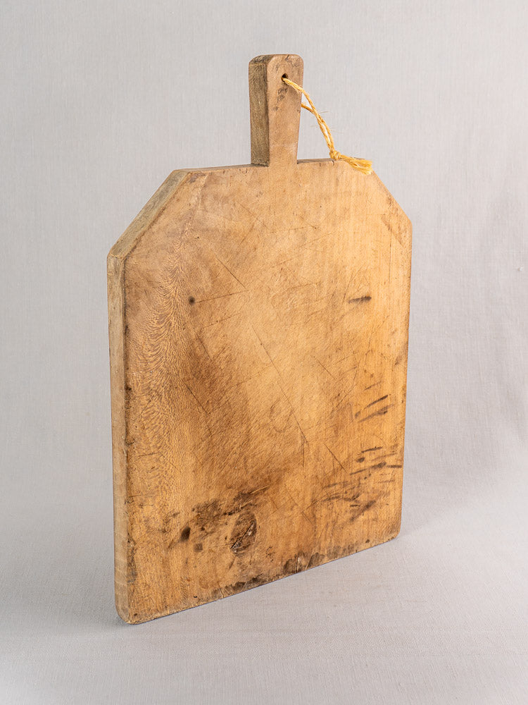 Antique French hand-carved beech bread board with handle and string, upright; vintage knife marks visible.
