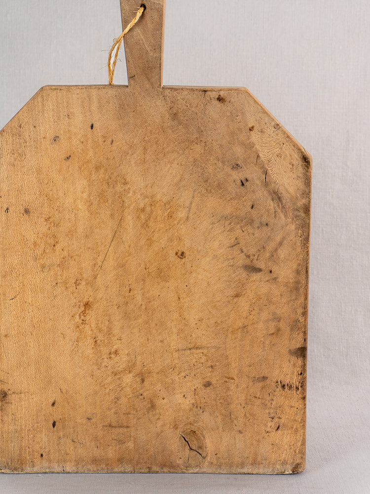 Antique French bread board in hand-carved beech, rectangular with handle and hanging string, light gray backdrop.