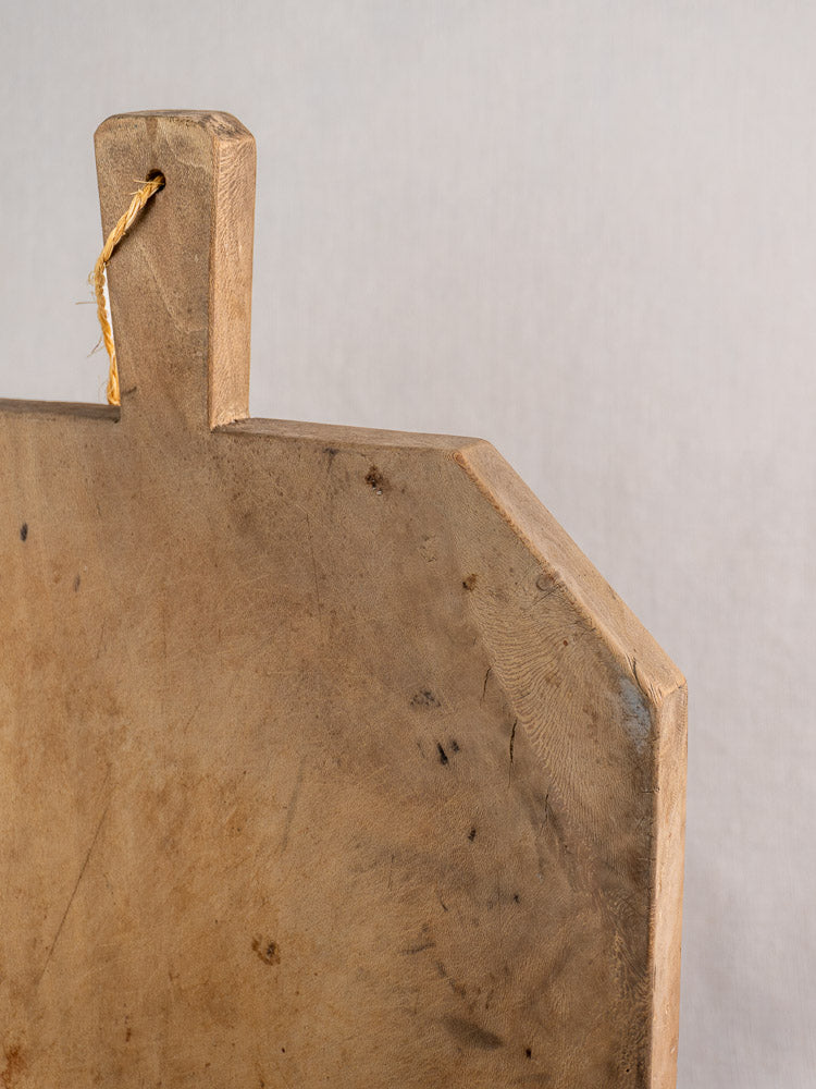 Top corner of antique French bread board in hand-carved beech, handle with string, neutral backdrop.