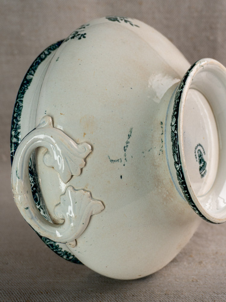 Upside-down antique French Saint-Amand Fruits Pattern faience soupière with ornate handles and green floral border, on beige textured surface.