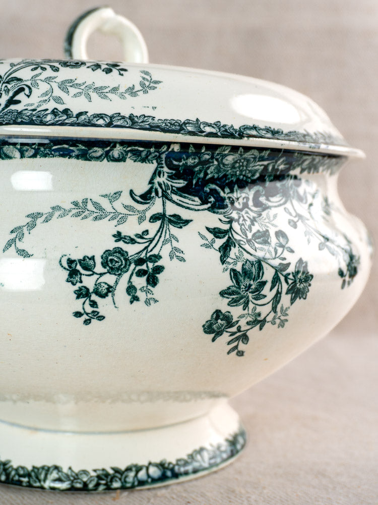 Antique French faience soup tureen with dark fruit and floral motifs, looped lid handle, on light fabric background.