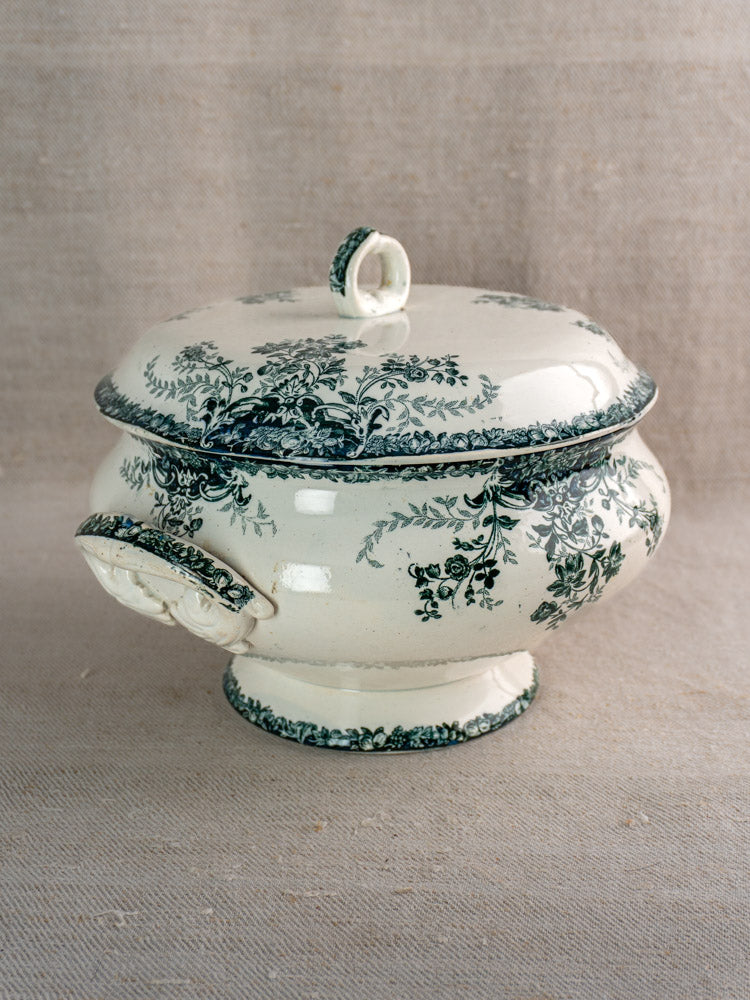 Antique French faience white ceramic soup tureen with dark green fruit motif and ornate lid, on beige fabric.