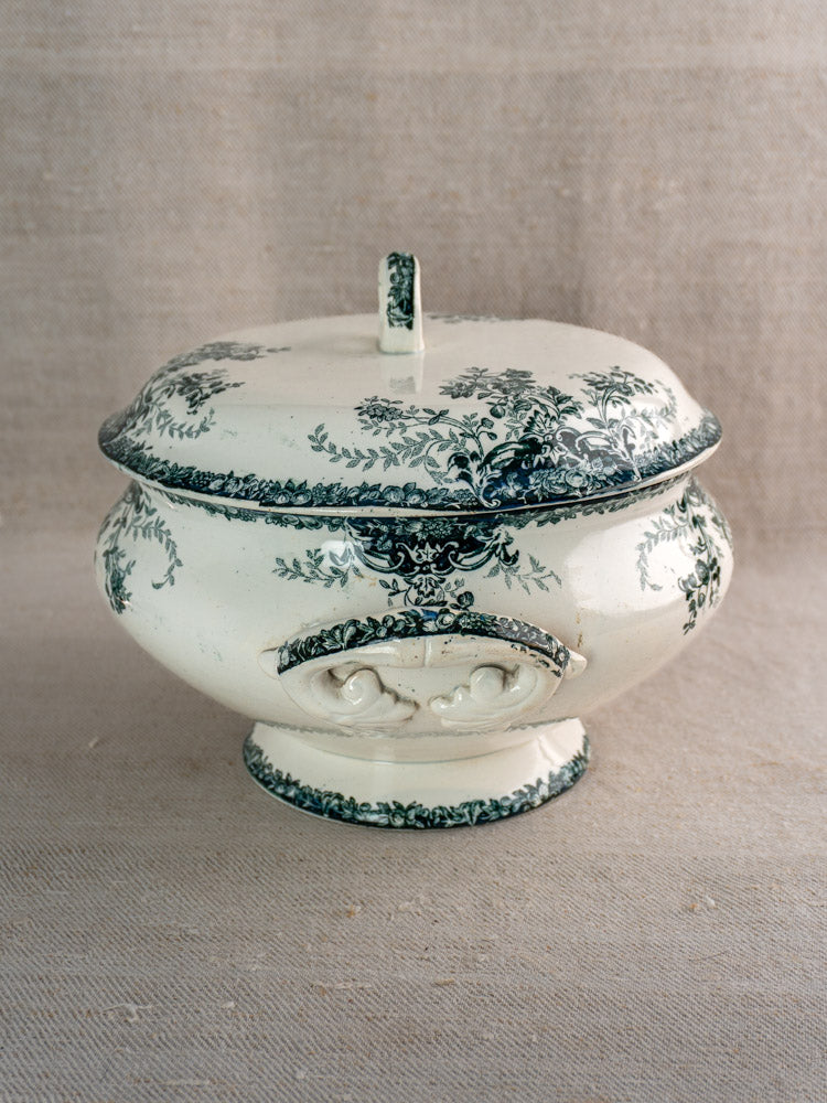 Antique French faience soup tureen with fruit motif on textured beige fabric background.