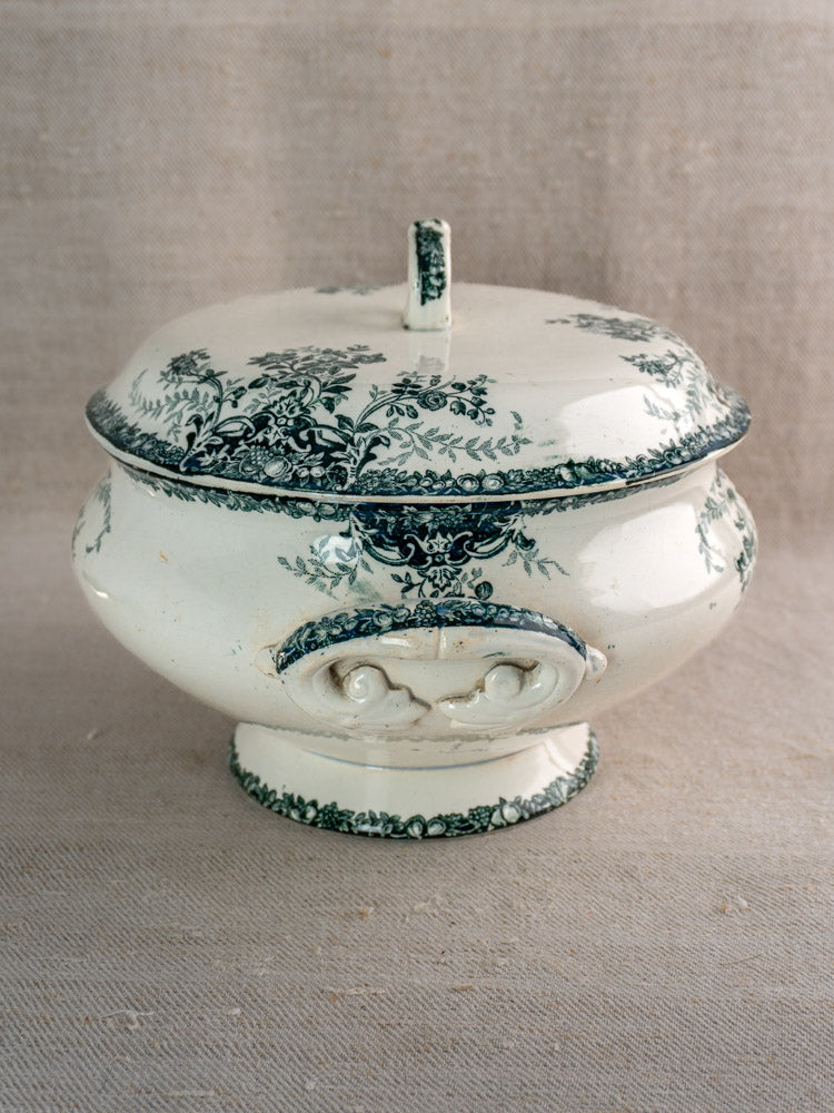 Antique French faience soup tureen with fruit pattern and ornate lid, on beige fabric surface.