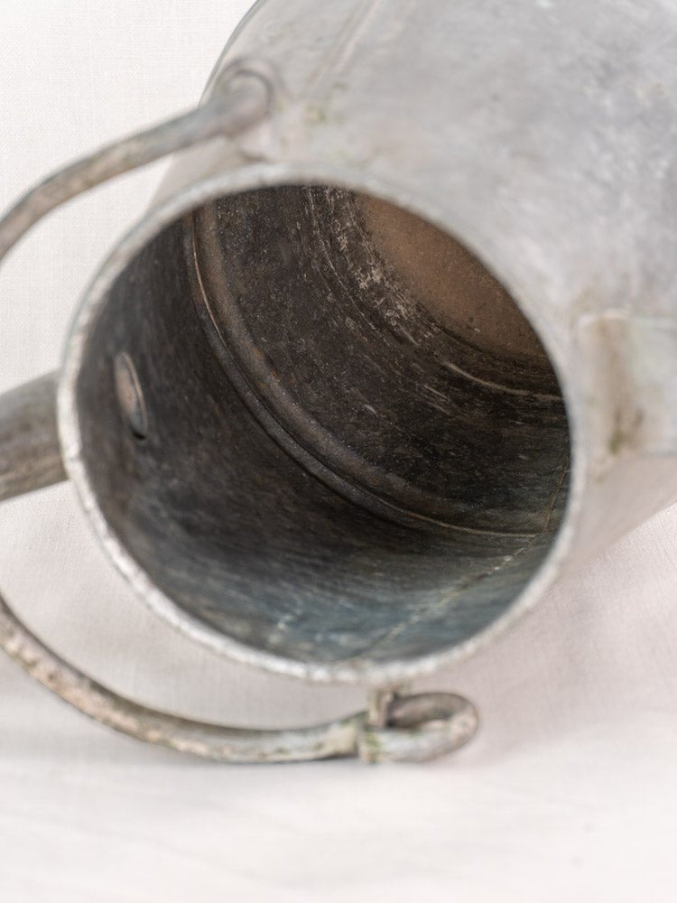 Antique French zinc watering can with milk can shape, spout, and handle on light fabric backdrop.