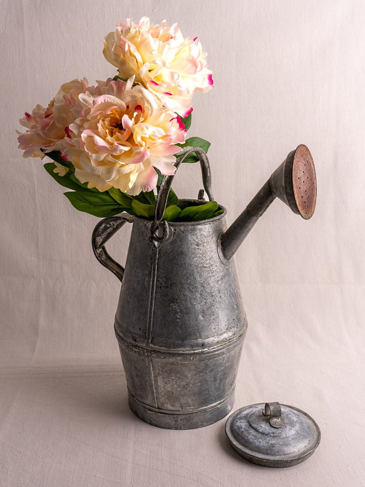 Antique French zinc watering can in classic milk can shape with peonies, lid on fabric, soft beige background.