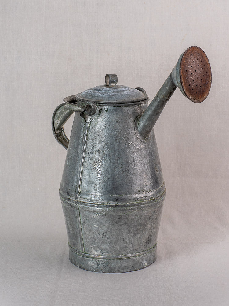 Antique French zinc watering can with milk can shape, side handle, domed lid, and long spout on a light fabric background.