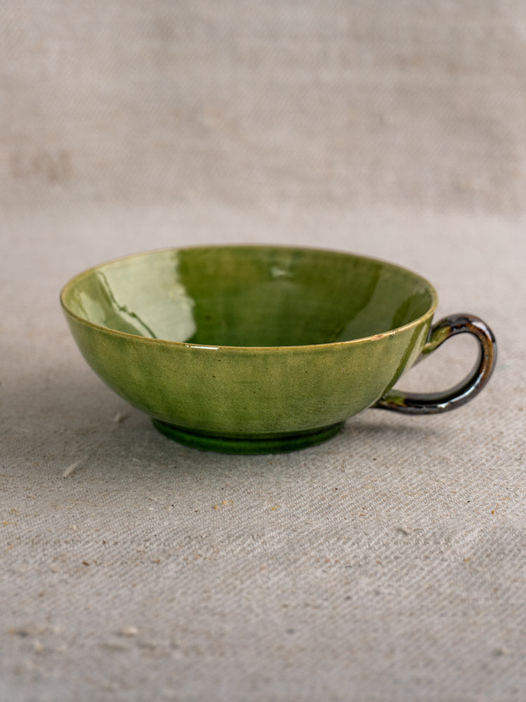 Dieulefit green glazed vintage French teacup with glossy finish and round handle on beige fabric background.