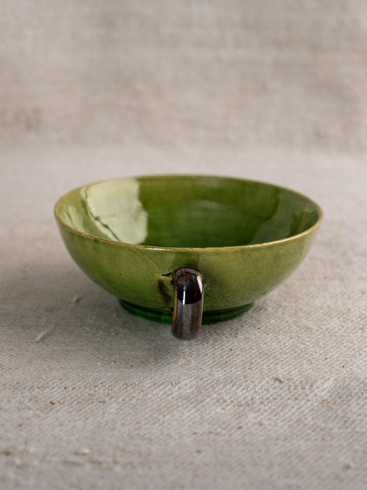 Vintage French Dieulefit green glaze teacup with shallow round shape and small dark handle on beige textured fabric.