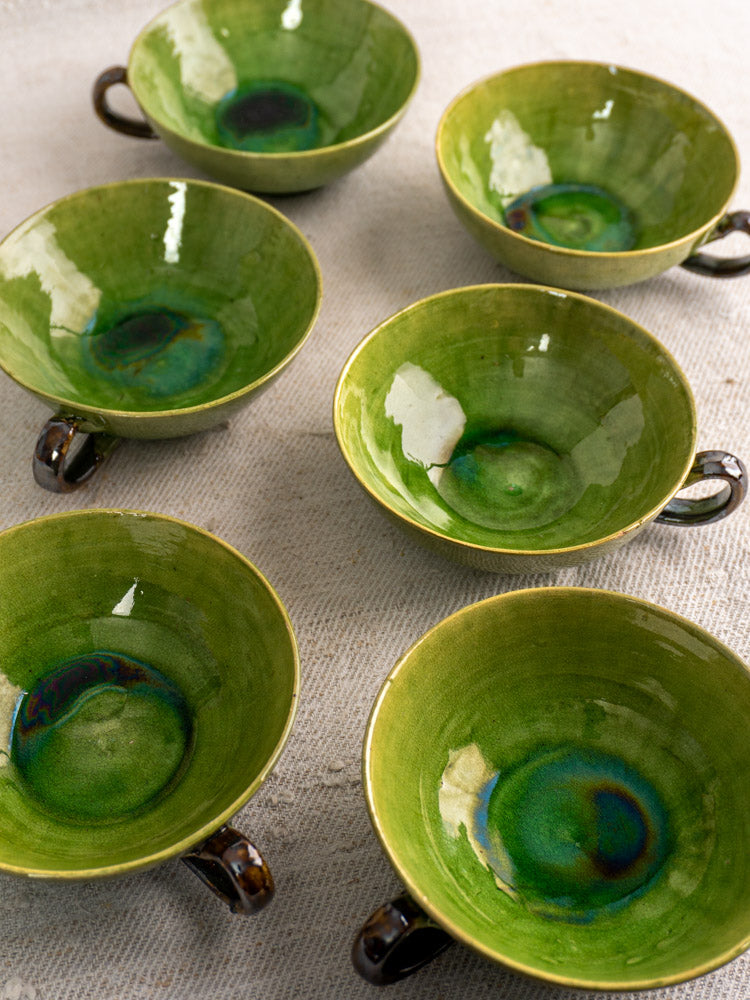 Six vintage French teacups with Dieulefit green glaze, blue-turquoise accents, and dark handles on a beige textured surface.