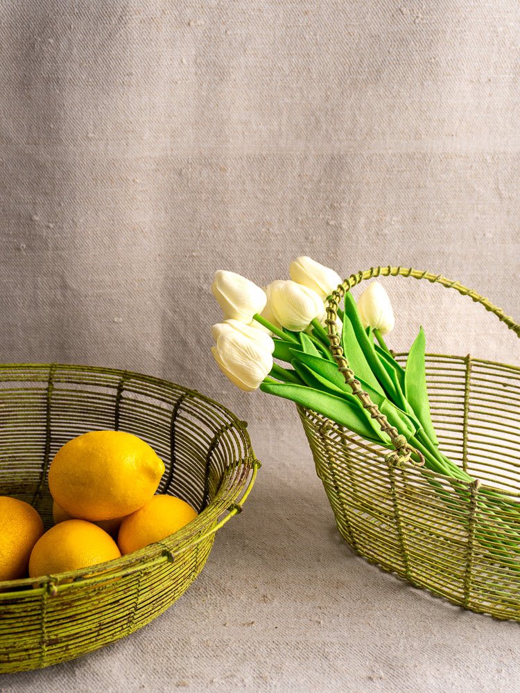 Antique French green fil de fer wire baskets, one with lemons, one with white tulips, on beige fabric.